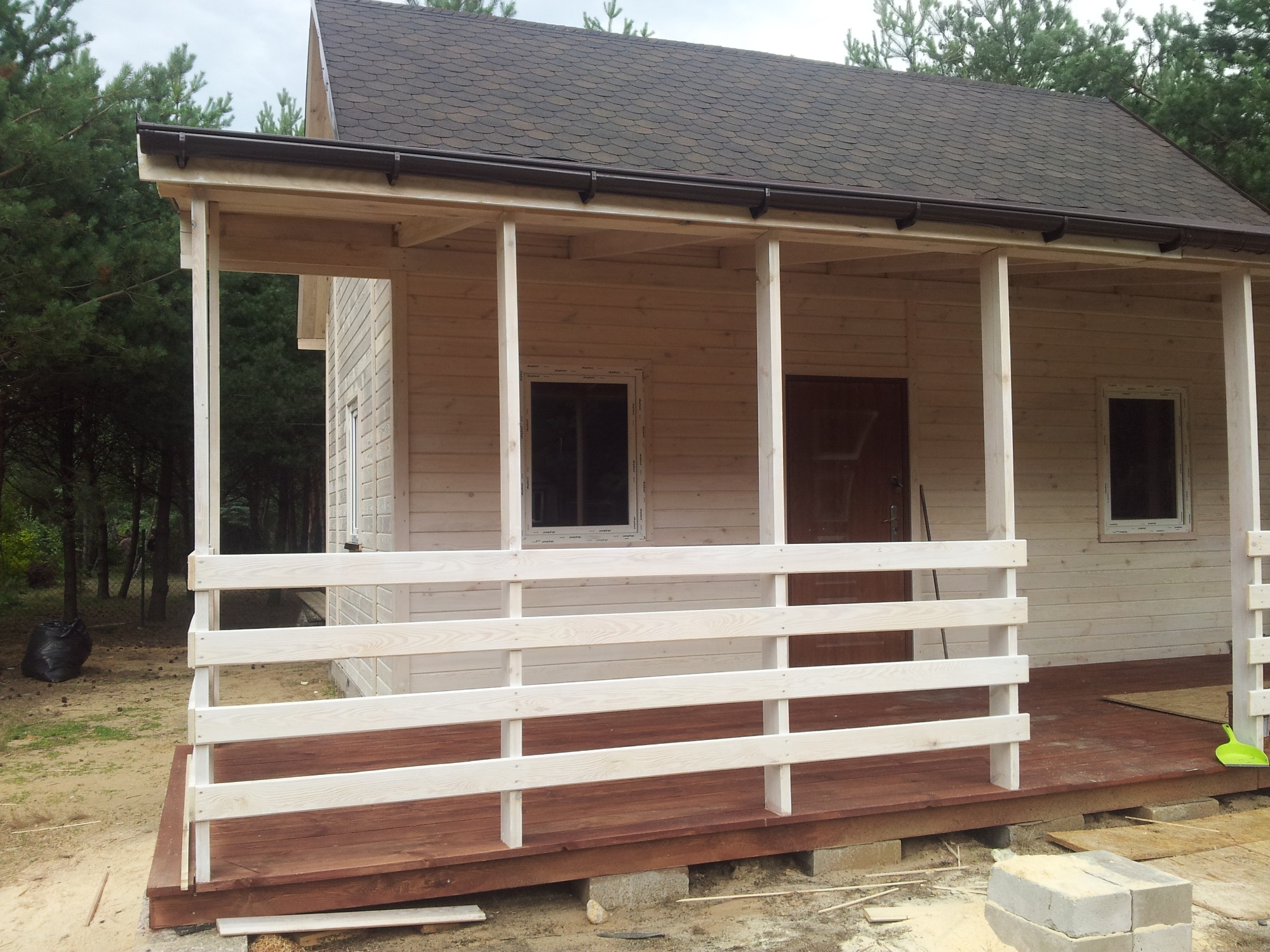 Surowy drewniany dom z gontowym dachem i zadaszonym gankiem z białą balustradą, w otoczeniu drzew iglastych.