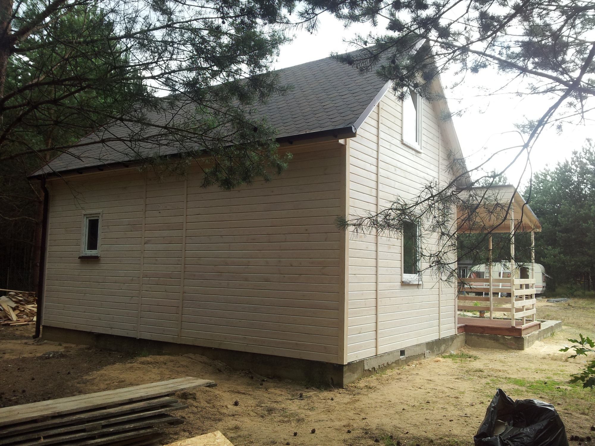 Surowy, drewniany dom z ciemnym dachem z gontów bitumicznych, otoczony sosnowym lasem, widoczne deski i worek na śmieci.