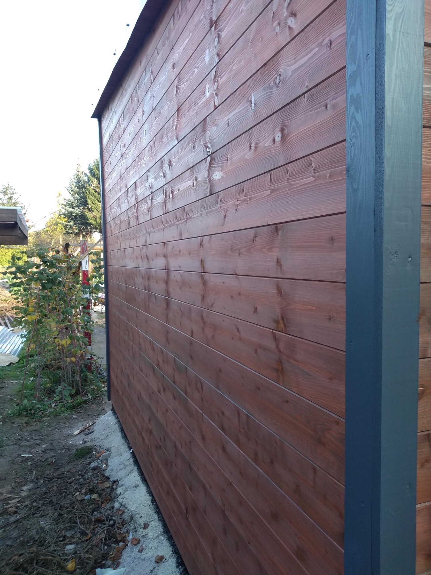 Ściana budynku z brązowych desek, z pomalowanym na szaro narożnikiem, widok z zewnątrz, z fragmentem roślinności w tle.