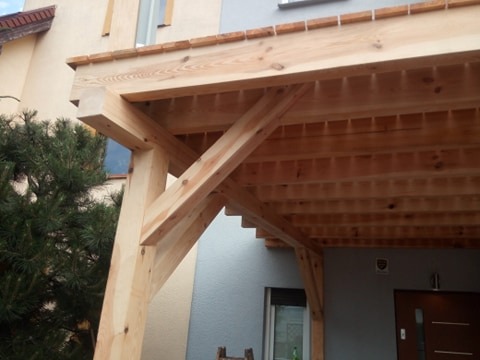 Drewniana konstrukcja z belek tworząca zadaszenie nad wejściem do domu, widoczne elementy dachu i wspierające słupy.