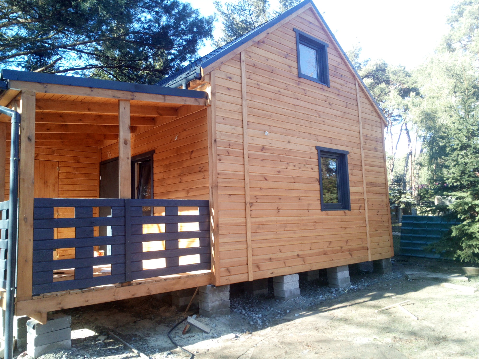 Niedokończony drewniany dom szkieletowy z ciemną stolarką okienną i zadaszonym tarasem z ażurową balustradą, posadowiony na betonowych bloczkach, widok z boku w słoneczny dzień.
