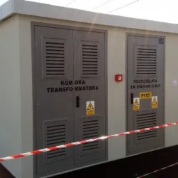 Zewnętrzna, betonowa stacja transformatorowa z dwoma szarymi, metalowymi drzwiami z kratkami wentylacyjnymi i tabliczkami ostrzegawczymi, otoczona taśmą ostrzegawczą.