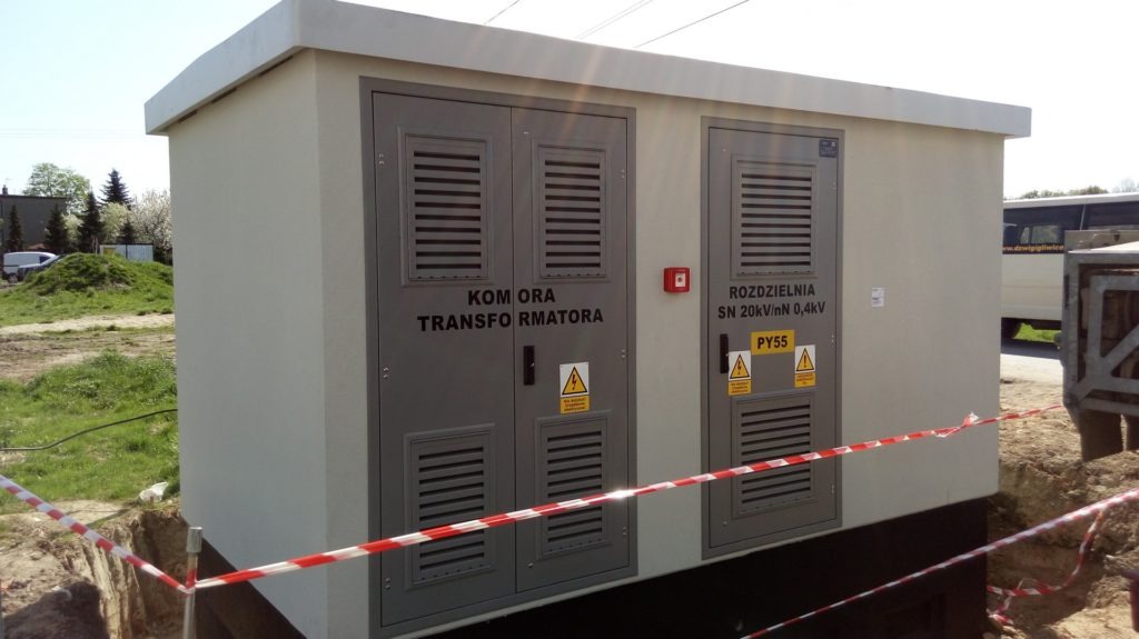 Zewnętrzna, betonowa stacja transformatorowa z dwoma szarymi, metalowymi drzwiami z kratkami wentylacyjnymi i tabliczkami ostrzegawczymi, otoczona taśmą ostrzegawczą.