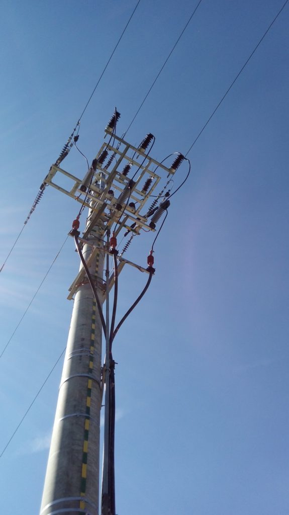 Betonowy słup energetyczny z zamontowaną konstrukcją stalową i izolatorami, widok z dołu na tle błękitnego nieba.