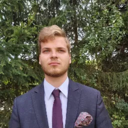 Młody mężczyzna w garniturze z poszetką, pozujący na tle zielonych drzew.