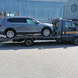Szary samochód osobowy marki Volkswagen Tiguan transportowany na lawecie pomocy drogowej przed salonem samochodowym marki Volkswagen.