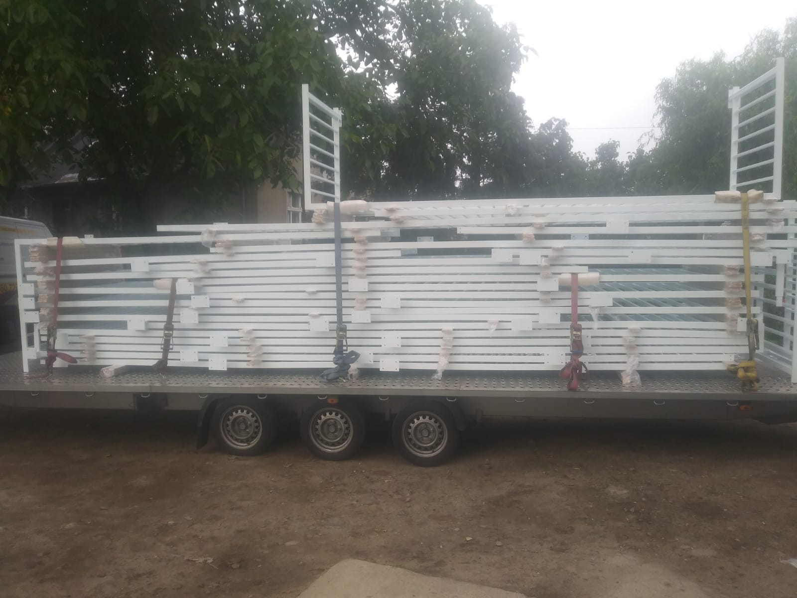 Przyczepa z ładunkiem białych, metalowych elementów ogrodzeniowych, zabezpieczonych pasami transportowymi, widoczne koła przyczepy i zieleń w tle.