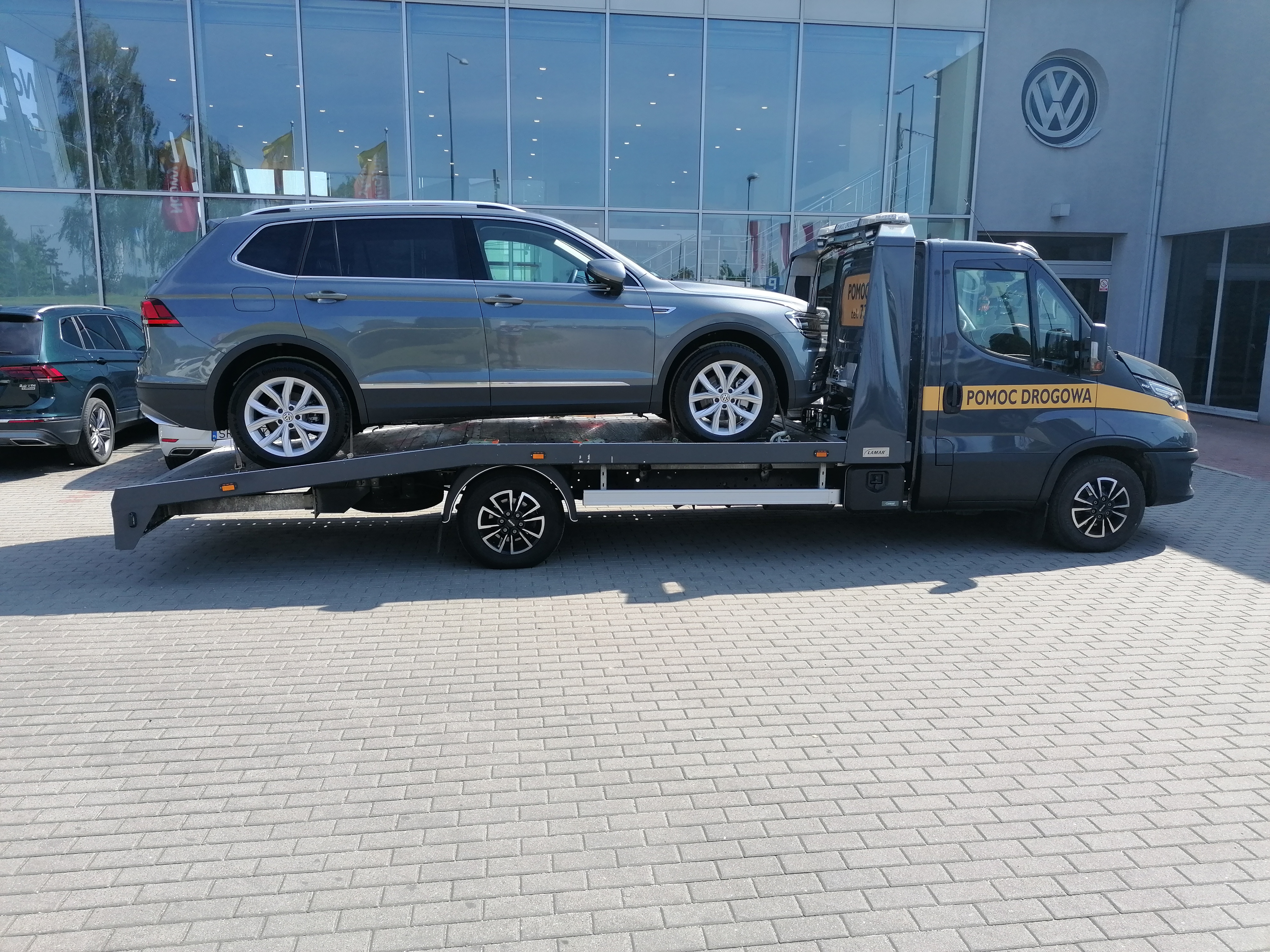Szary samochód osobowy marki Volkswagen Tiguan transportowany na lawecie pomocy drogowej przed salonem samochodowym marki Volkswagen.