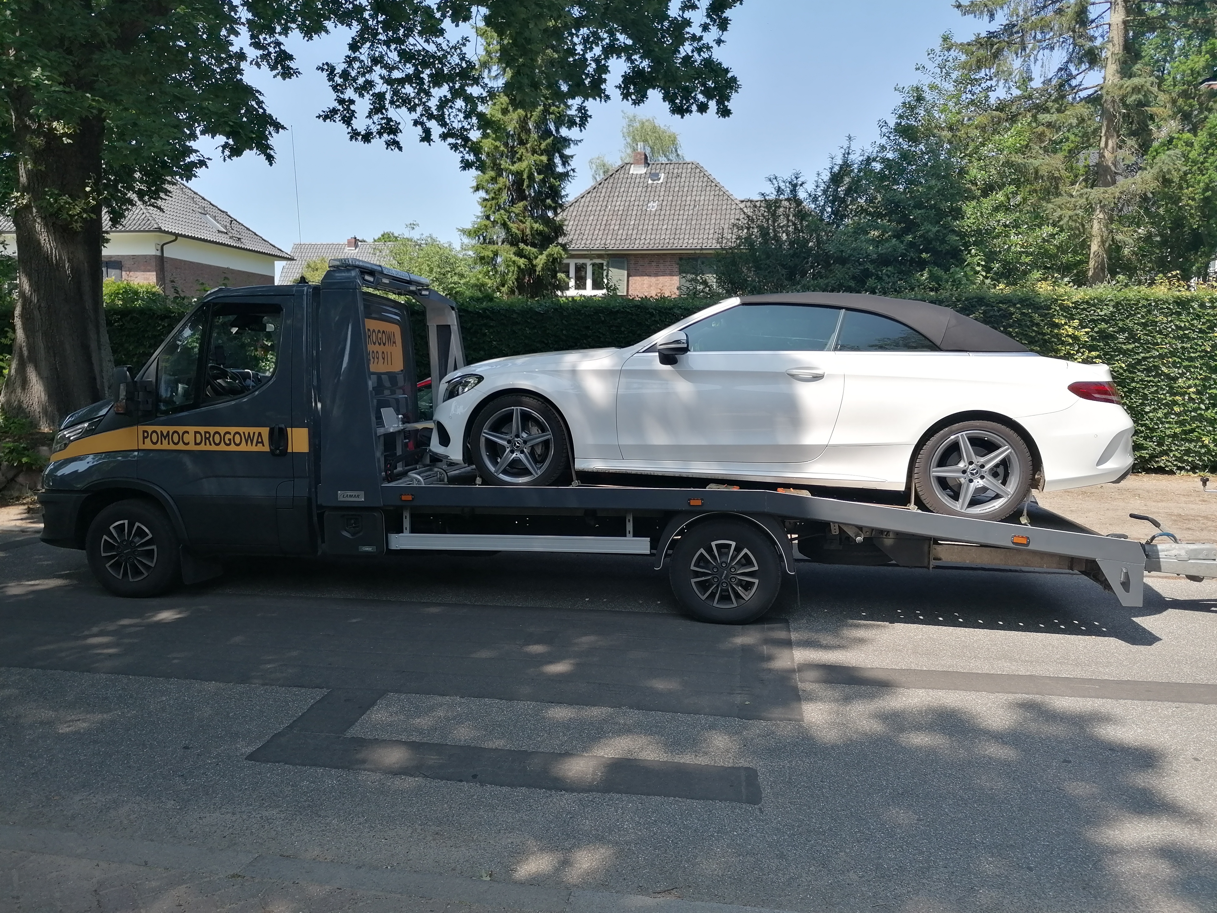 Laweta z napisem 'Pomoc Drogowa' przewozi białego Mercedesa kabrioleta na tle zieleni i domów.