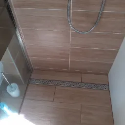 Ukończona kabina prysznicowa z odpływem liniowym w podłodze, wykończona płytkami imitującymi drewno, widoczny wąż prysznicowy i akcesoria toaletowe za szybą.