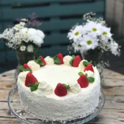 Tort kokosowy udekorowany truskawkami, bitą śmietaną i miętą, podany na szklanej paterze na drewnianym stole, z bukietami białych kwiatów w tle.