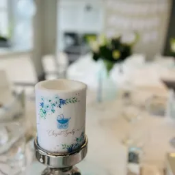 Świeca dekoracyjna z motywem wózka dziecięcego i napisem 'Chrzest Święty' na srebrnym świeczniku, ustawiona na stole z zastawą podczas uroczystości.