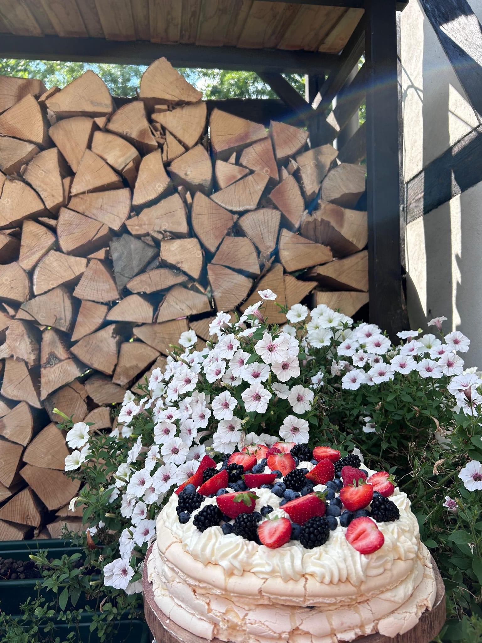 Tort beza udekorowany świeżymi truskawkami, jeżynami i borówkami, ustawiony na drewnianym podeście na tle białych kwiatów i stosu drewna opałowego.