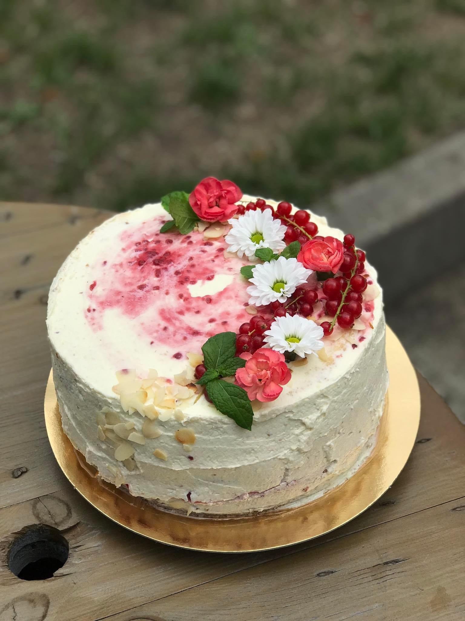 Okrągły tort z białym kremem, dekorowany czerwonymi porzeczkami, różowymi i białymi kwiatami oraz listkami mięty, umieszczony na złotej podkładce na drewnianym stole.