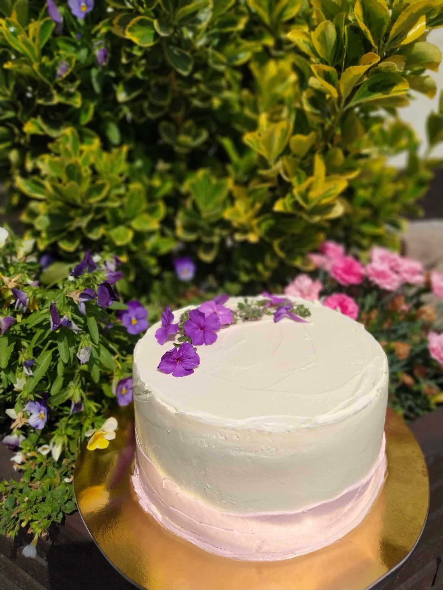 Tort z białym kremem i fioletowymi kwiatami na złotej podkładce, otoczony zielenią i różowymi kwiatami, sfotografowany w naturalnym świetle.