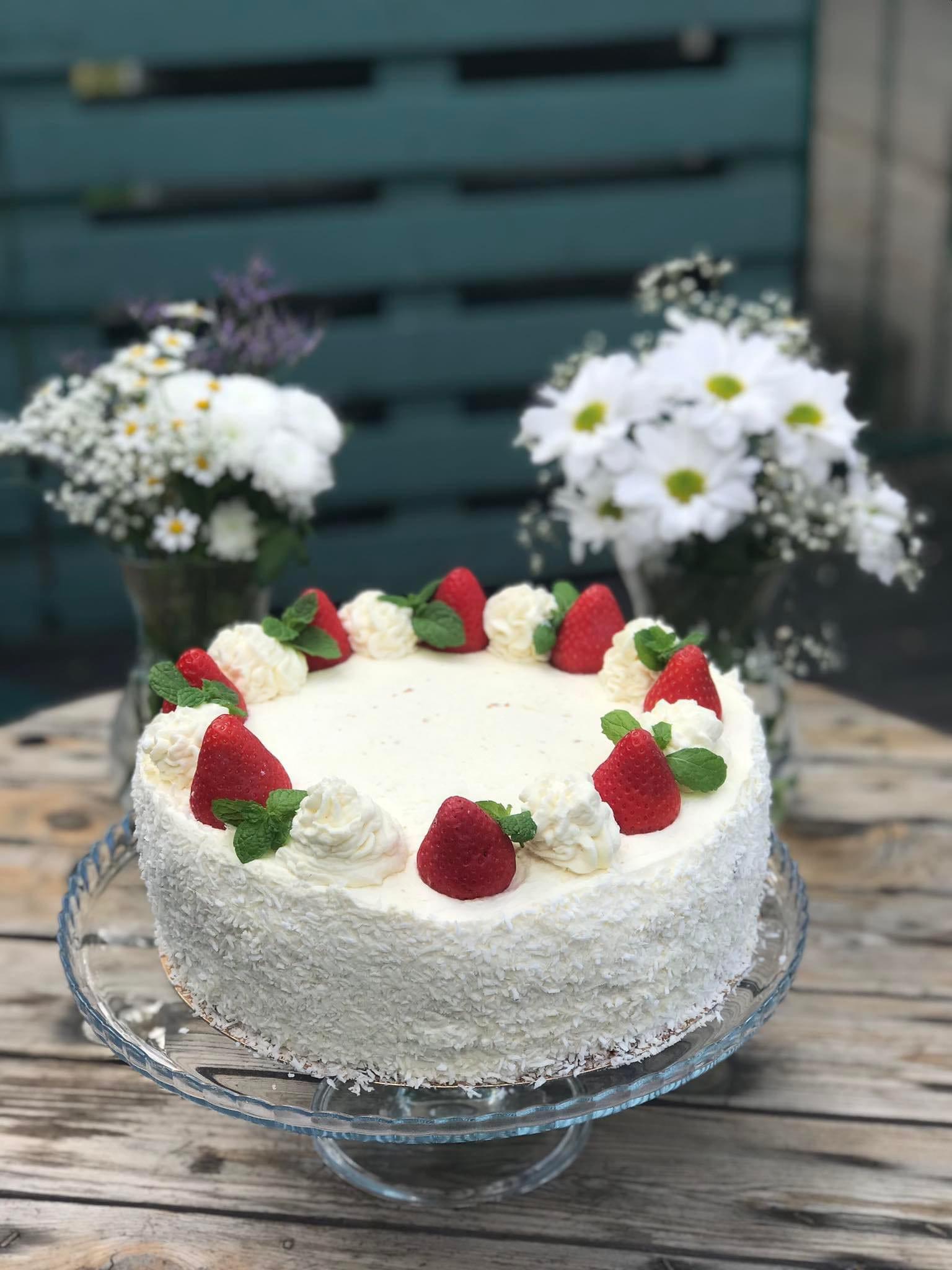 Tort kokosowy udekorowany truskawkami, bitą śmietaną i miętą, podany na szklanej paterze na drewnianym stole, z bukietami białych kwiatów w tle.