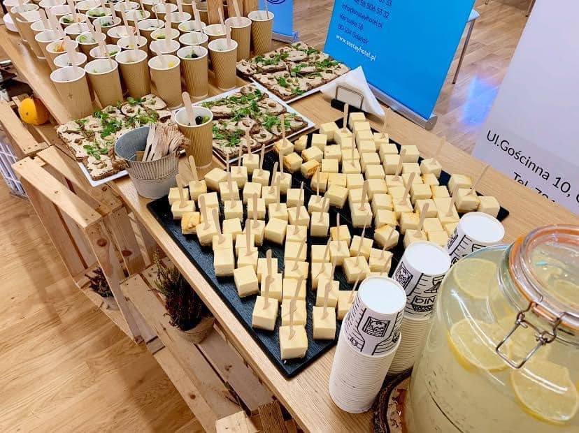 Stół cateringowy z przekąskami: kostki sera na czarnym talerzu z wykałaczkami, kanapki z zielonymi dodatkami, napoje w kubkach papierowych i lemoniada w dużym słoju z cytryną, ustawione...