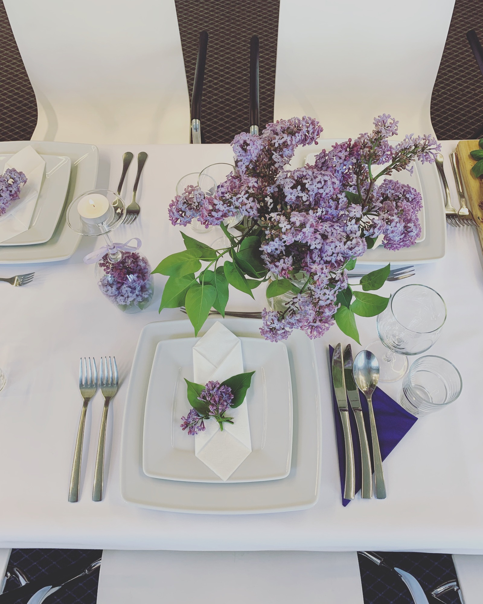 Elegancko nakryty stół z białym obrusem, kwadratowymi talerzami, srebrnymi sztućcami i bukietem bzów w szklanym wazonie; dodatkowo świeczka w szklanym świeczniku i dekoracje z bzu na serwetkach.