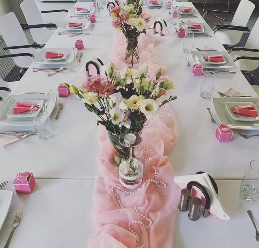 Uroczysty stół z białym obrusem, różową organzą, kwiatami w wazonach, elegancką zastawą, szklankami, przyprawnikami i małymi różowymi pudełeczkami.