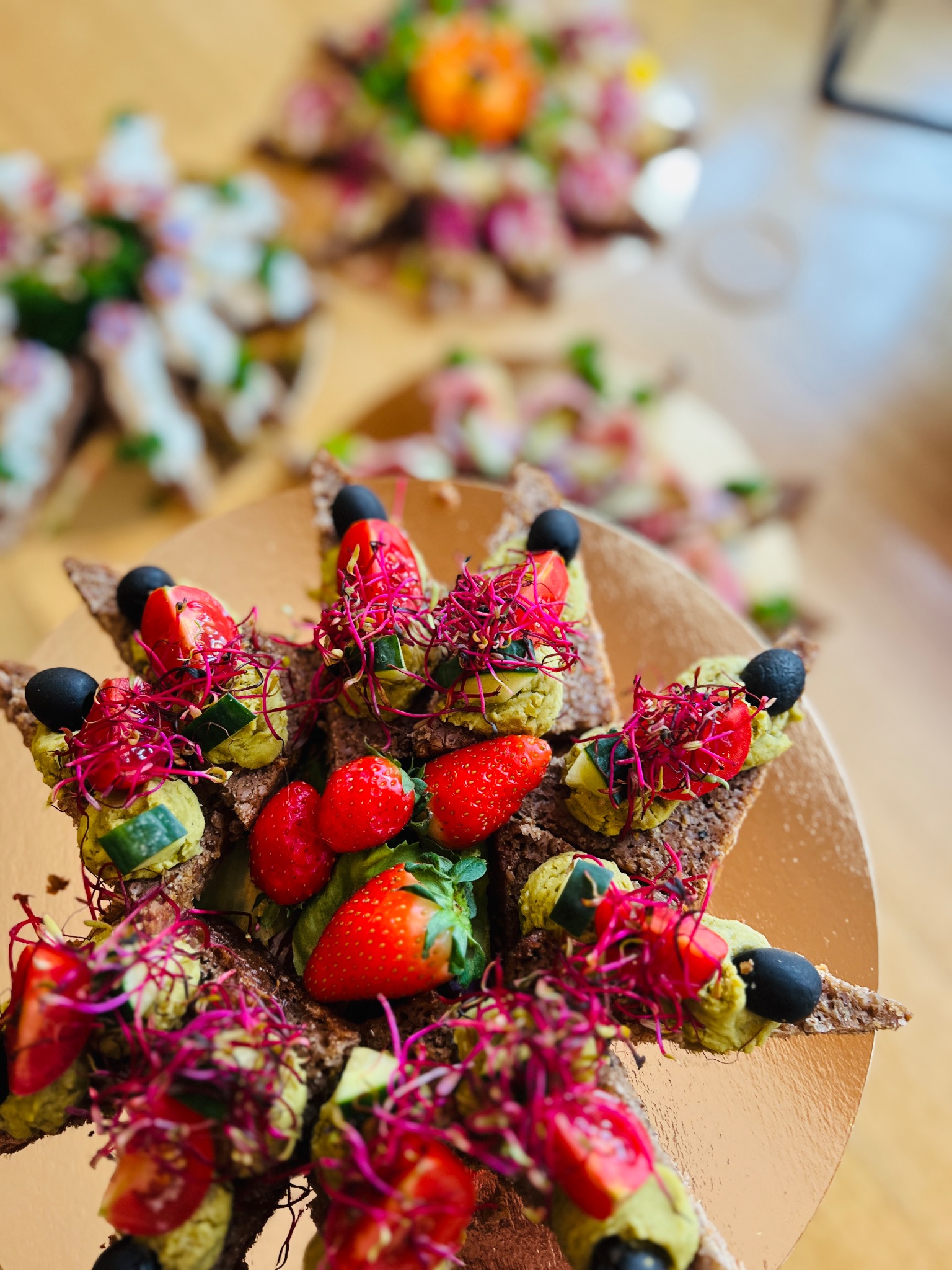 Starannie ułożone na złotej tacy kanapki na ciemnym pieczywie z pastą, pomidorkami, ogórkiem, oliwkami i czerwoną rzodkiewką, tworzące kompozycję z truskawkami w centrum.