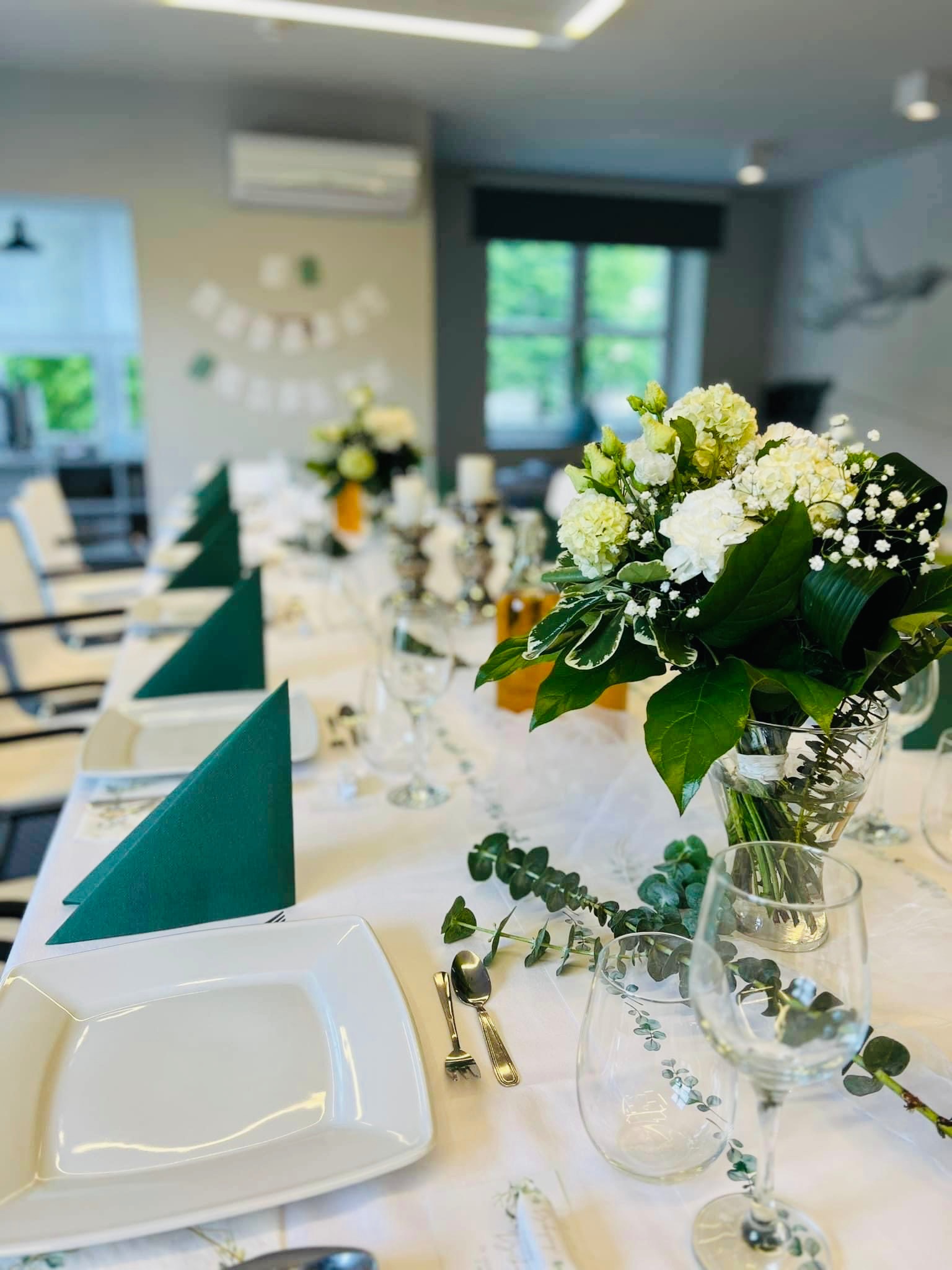 Uroczysty stół komunijny: biały obrus, kwadratowe talerze, ciemnozielone serwetki złożone w trójkąt, bukiety z białych kwiatów w szklanych wazonach, sztućce i kieliszki z motywem eukaliptusa...