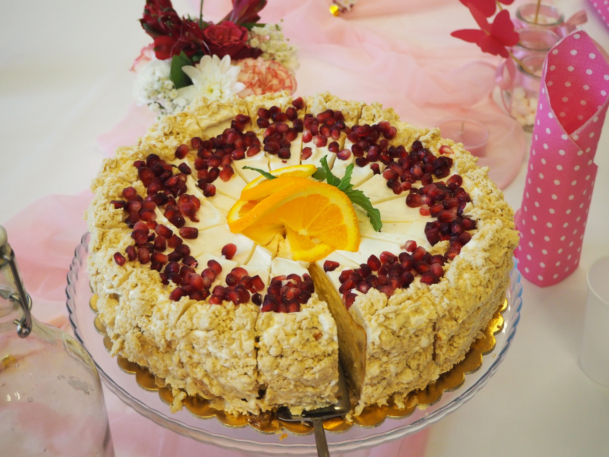 Kremowy tort z kruszonką, dekorowany granatem i pomarańczą, na szklanej paterze ze złotym brzegiem, z wyciętym kawałkiem, oraz kwiaty i dekoracje w tle.