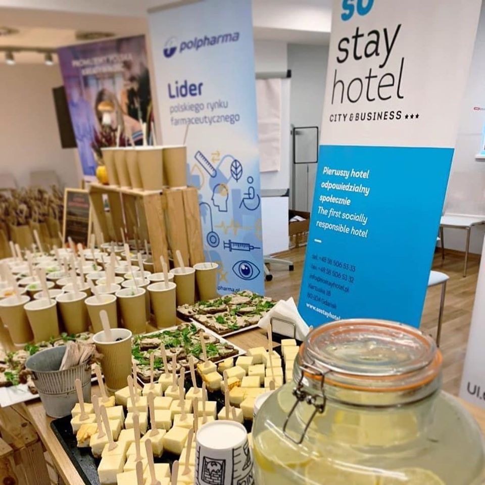 Stół z przekąskami: koreczki serowe na drewnianych wykałaczkach, kanapki z ziołami, napoje w papierowych kubkach, duży słój z wodą i cytryną, w tle banery reklamowe firm Polpharma i Stay Hotel.