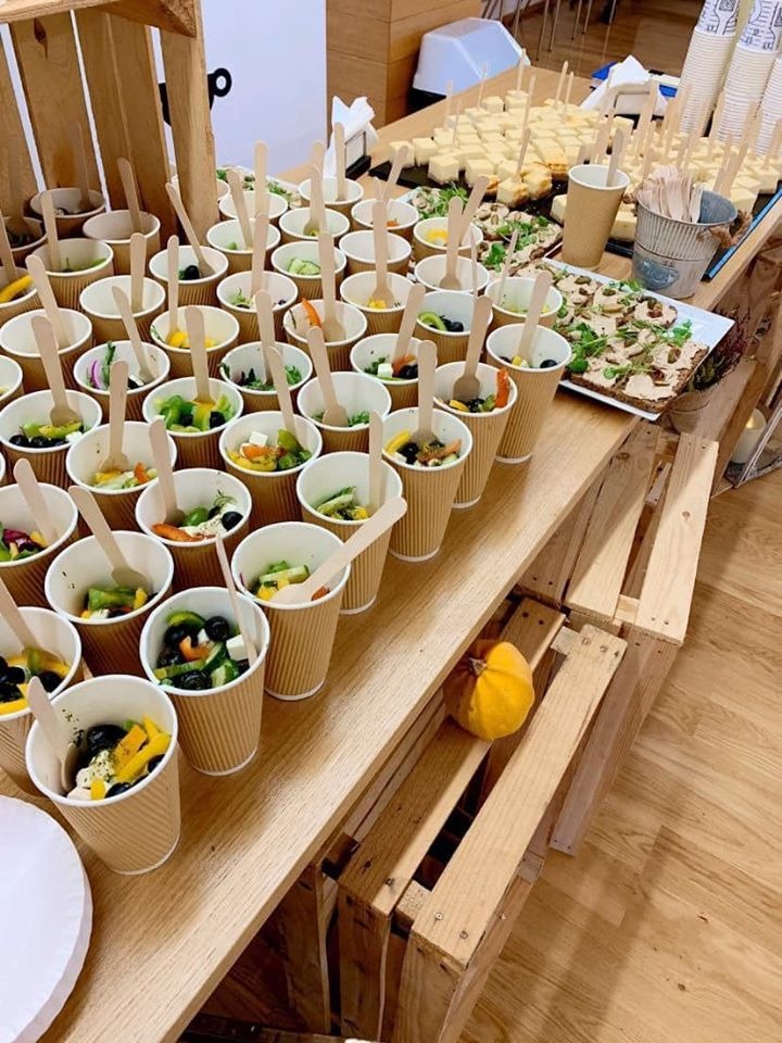 Rządki papierowych kubeczków z sałatkami i drewnianymi łyżeczkami na drewnianym stole, obok taca z przekąskami i mała dynia.