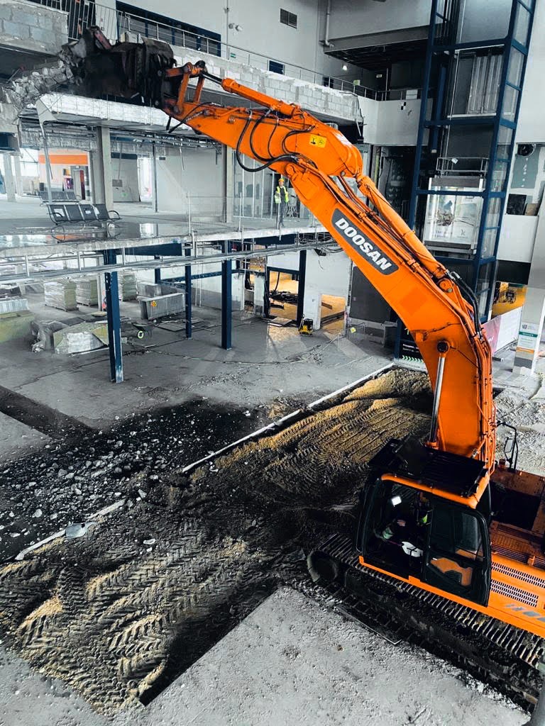 Pomarańczowa koparka Doosan kruszy sufit wewnątrz budynku podczas prac rozbiórkowych, widoczny operator w kabinie i pracownik w kamizelce odblaskowej na wyższym poziomie.