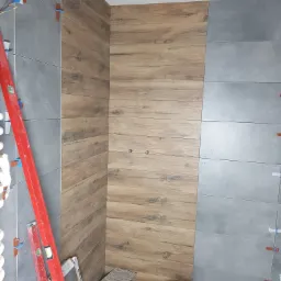 Ściana w trakcie wykańczania z szarych i drewnopodobnych płytek, widoczne poziomice, krzyżyki dystansowe i wiaderka z zaprawą.