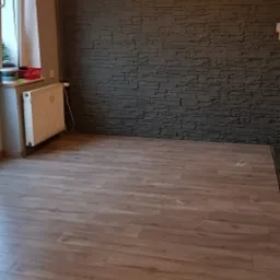 Jasne panele podłogowe ułożone w pustym pokoju z grzejnikiem i ścianą wyłożoną ciemnoszarymi płytkami imitującymi kamień.