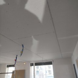 Gromix - Montaż płyt gipsowo-kartonowych na suficie, widoczne kable elektryczne, rusztowanie i podnośnik do płyt, cień osoby na suficie.
