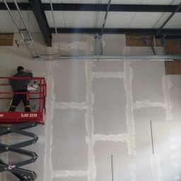Gromix - Pracownik na podnośniku montuje płyty gipsowo-kartonowe na ścianie w dużym pomieszczeniu; widoczne metalowe profile, instalacje pod sufitem i ślady szpachlowania łączeń płyt.
