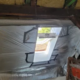 Folia termoizolacyjna zamocowana czarną taśmą wokół okna dachowego na poddaszu, widoczne drewniane belki konstrukcyjne i drabina.