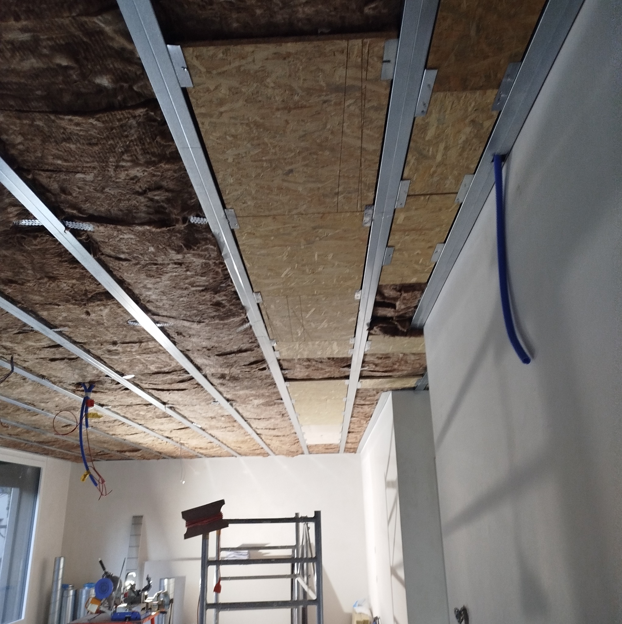 Wnętrze w trakcie prac: widok na sufit z widoczną izolacją z wełny mineralnej, płytami OSB i metalowym stelażem, fragment instalacji elektrycznej i drabina w tle.