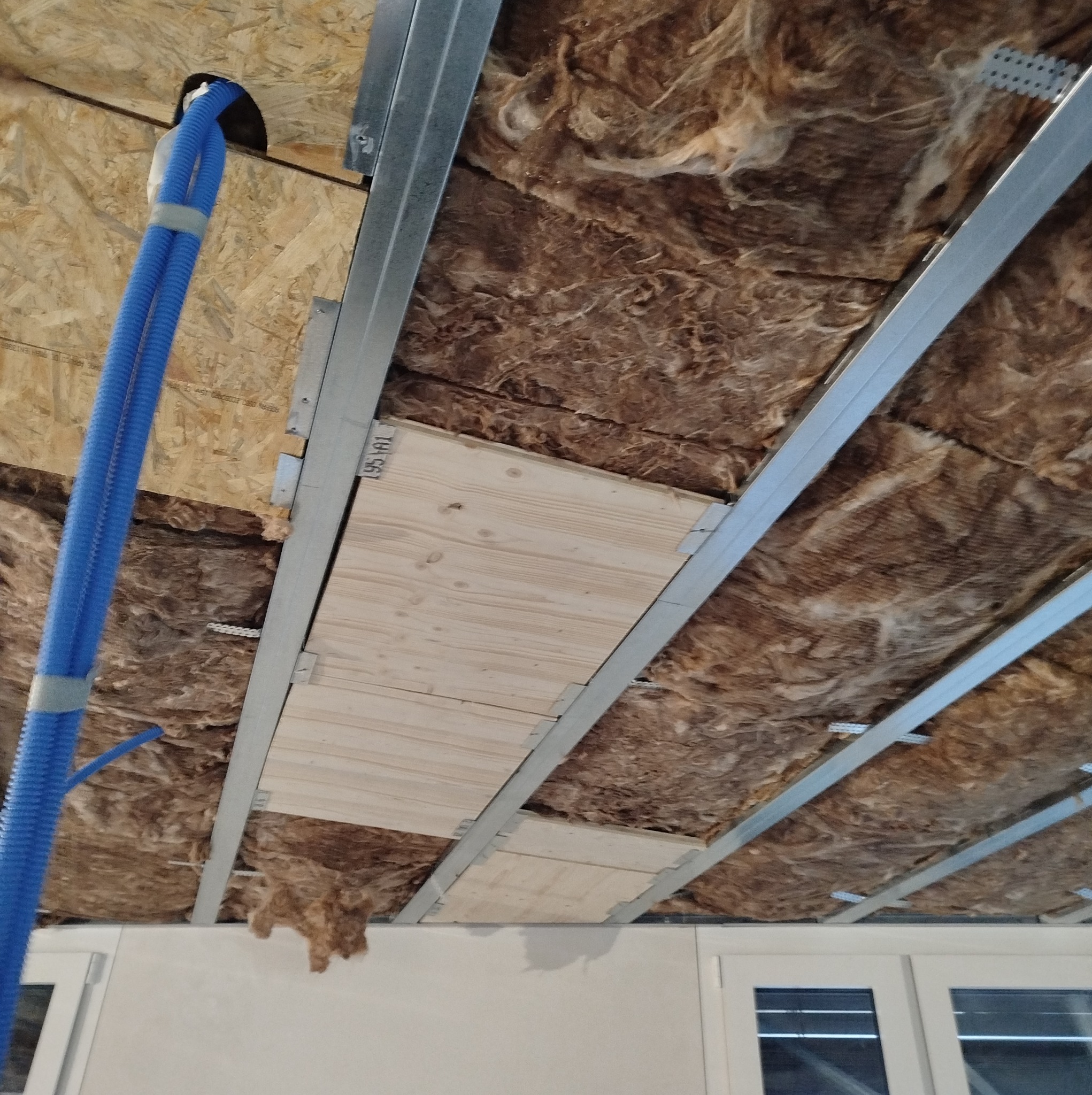 Poddasze w trakcie ocieplania: wełna mineralna między metalowymi profilami, częściowo zakryta drewnianymi płytami, widoczne niebieskie rury instalacyjne.