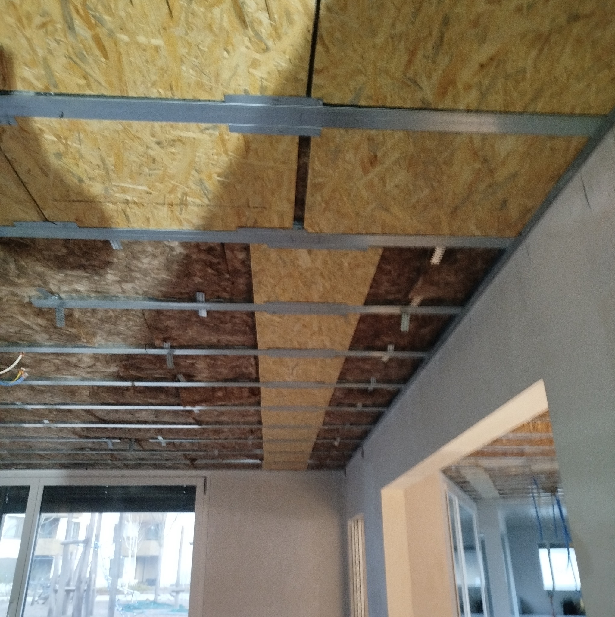 Montaż płyt OSB na metalowym ruszcie sufitowym, widoczne przewody elektryczne, surowy stan wykończenia wnętrza.