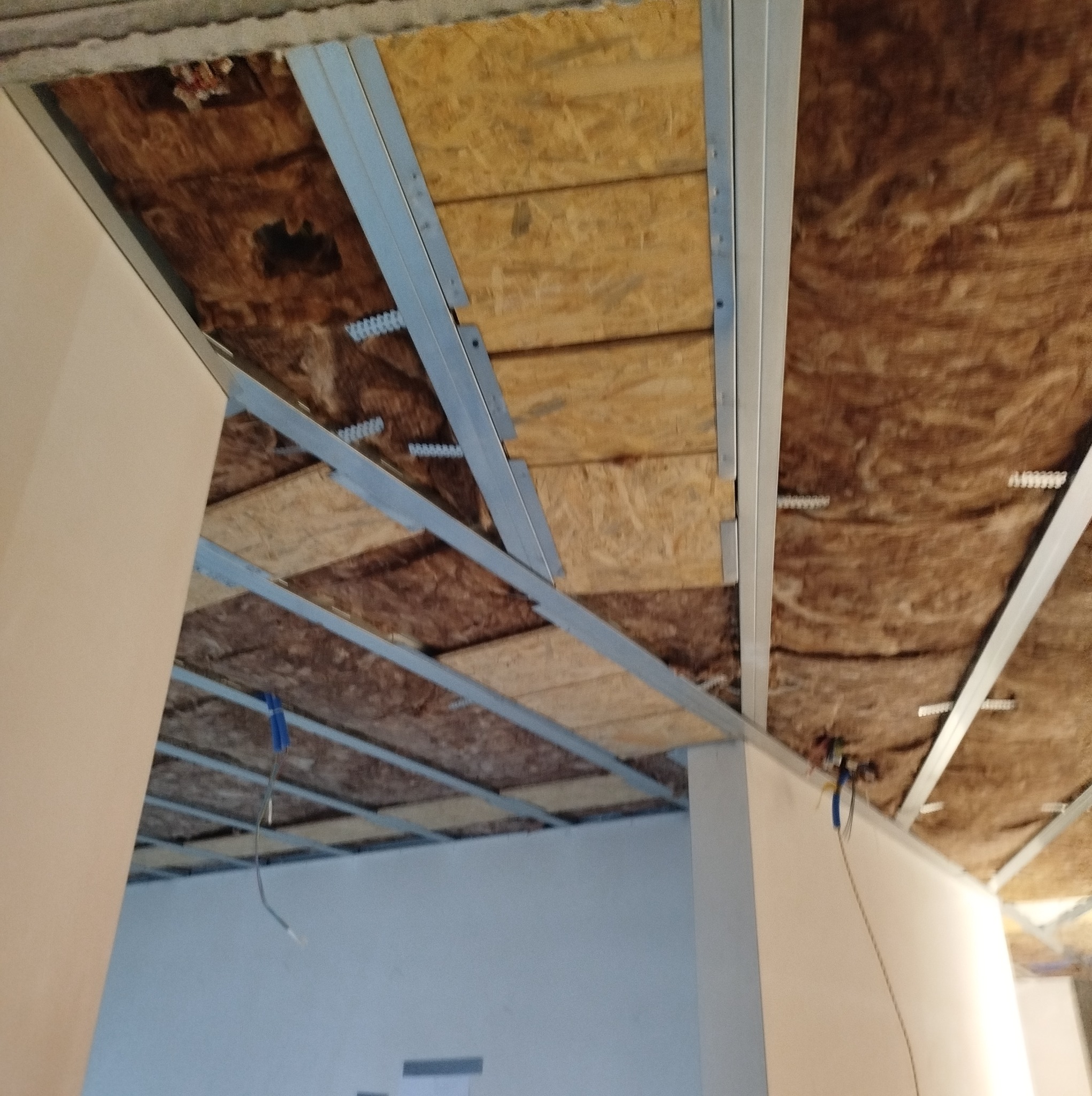 Konstrukcja metalowa sufitu podwieszanego z widoczną izolacją z wełny mineralnej i płytami OSB, zamontowana w pomieszczeniu z białymi ścianami.