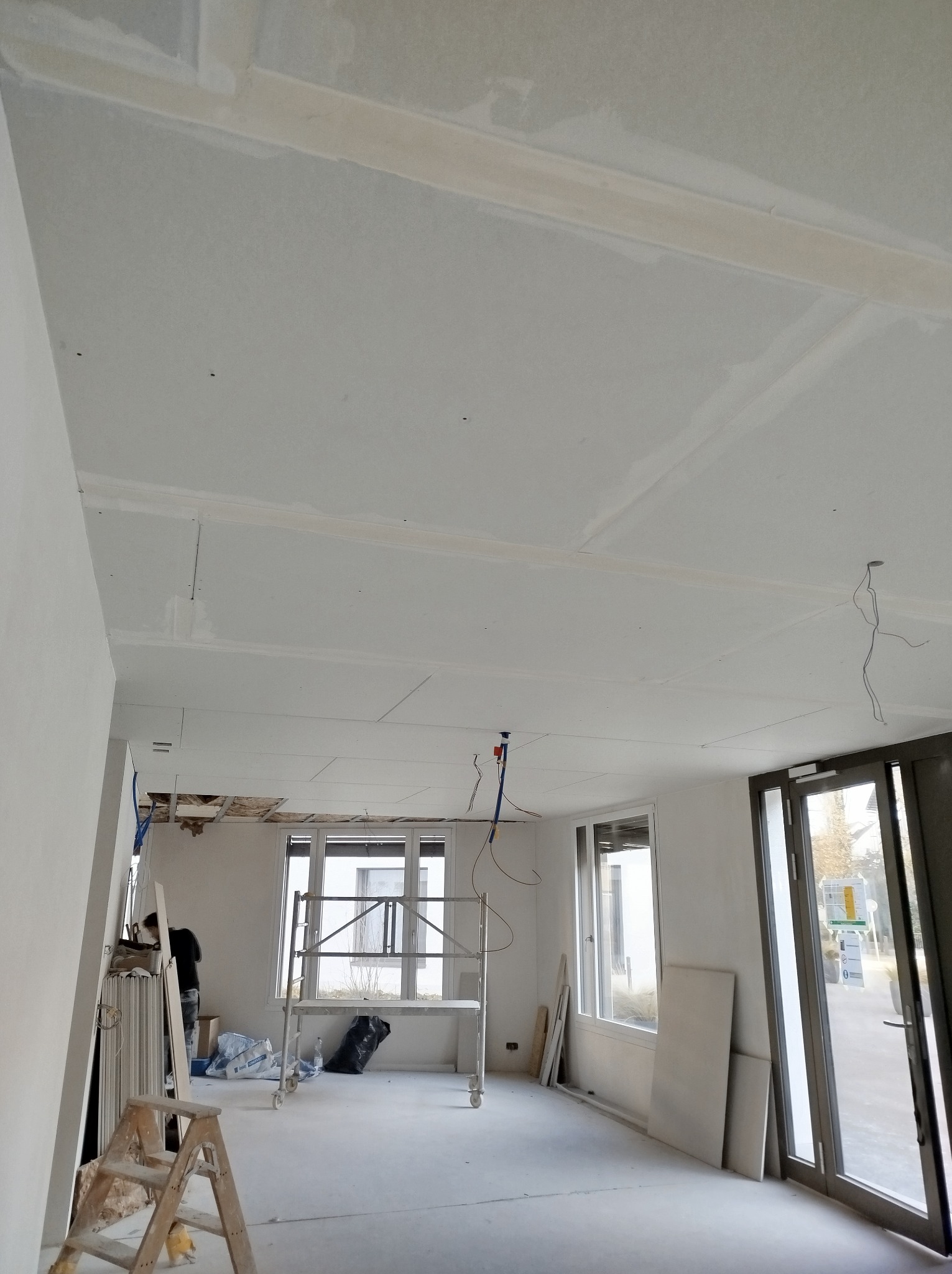 Wnętrze w trakcie prac wykończeniowych: sufit z płyt gipsowo-kartonowych z widocznymi łączeniami szpachlowanymi, częściowo odsłonięta konstrukcja stropu, drabina i rusztowanie w pomieszczeniu...