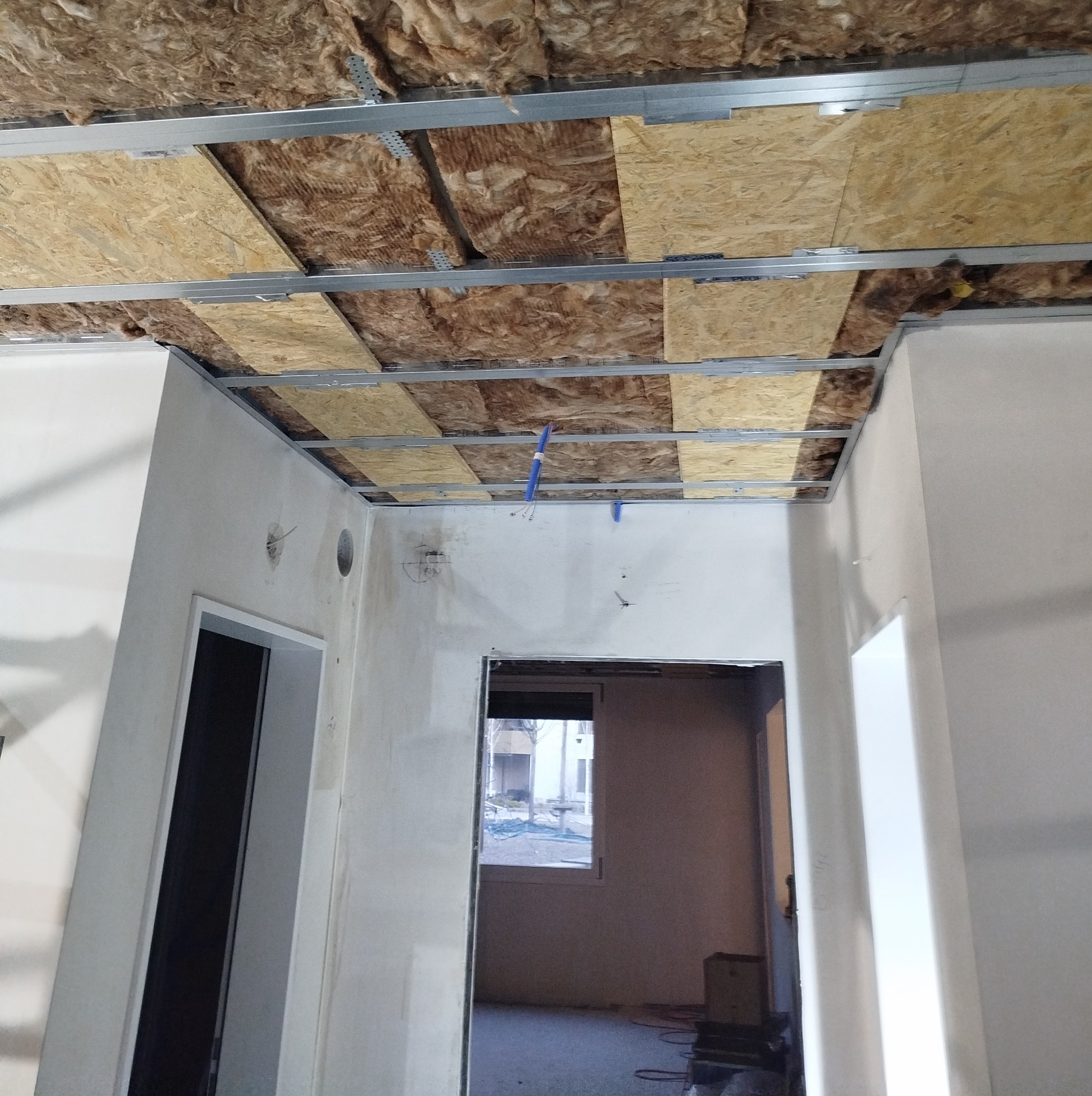 Wnętrze w trakcie prac wykończeniowych: widoczna konstrukcja metalowa podwieszanego sufitu z izolacją z wełny mineralnej i płytami OSB, przygotowana do dalszych prac, z widocznymi przewodami...