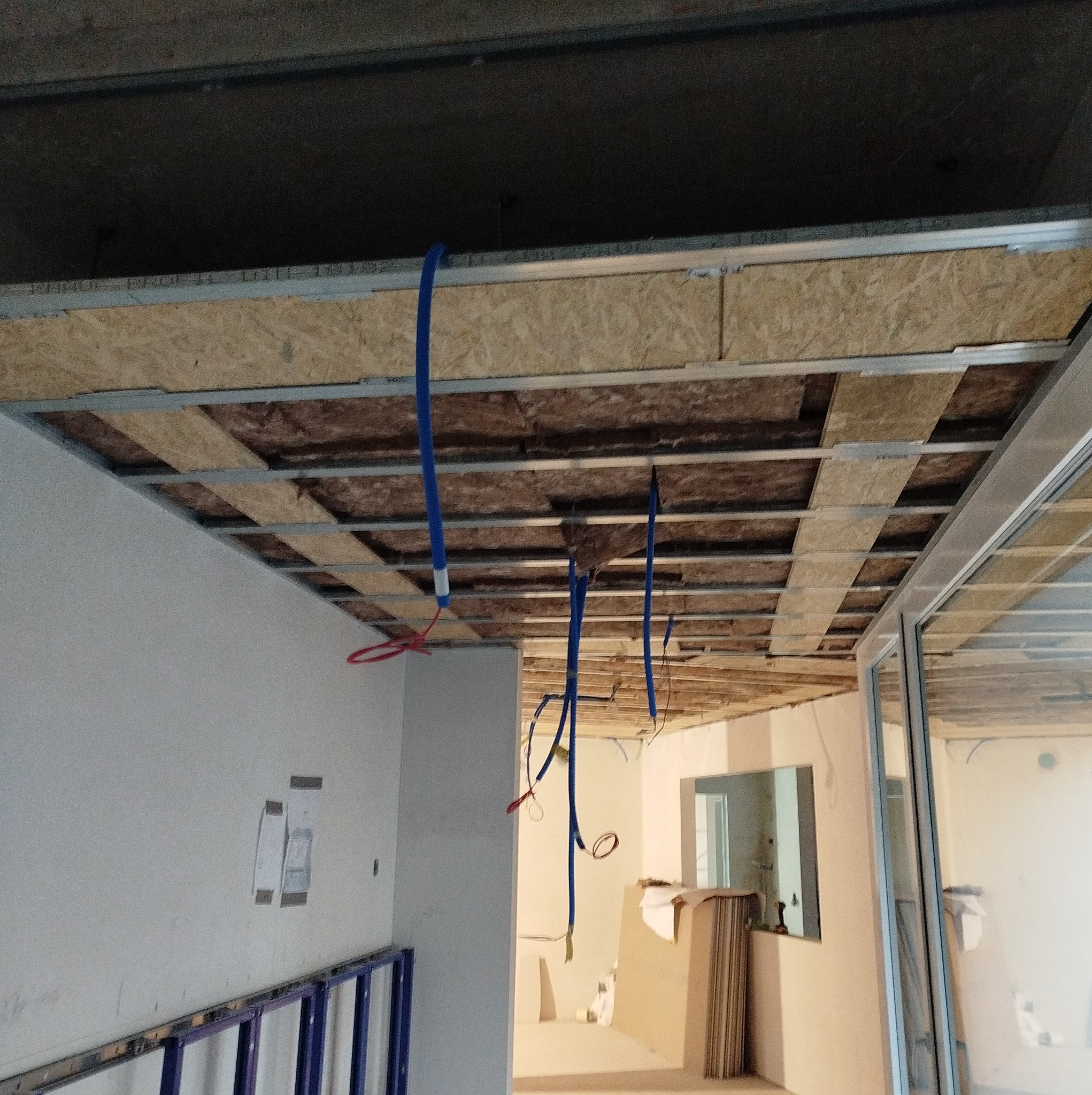 Montaż sufitu podwieszanego: widoczne profile metalowe, izolacja z wełny mineralnej, płyty OSB oraz zwisające kable instalacji elektrycznej.