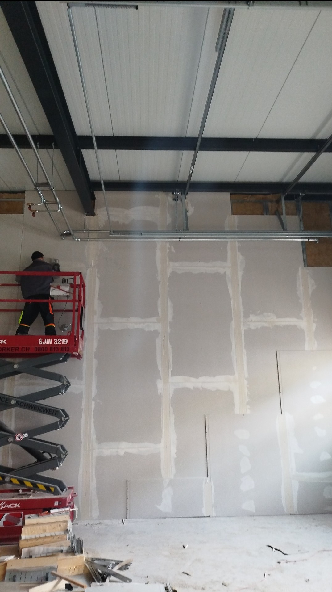 Pracownik na podnośniku montuje płyty gipsowo-kartonowe na ścianie w dużym pomieszczeniu; widoczne metalowe profile, instalacje pod sufitem i ślady szpachlowania łączeń płyt.