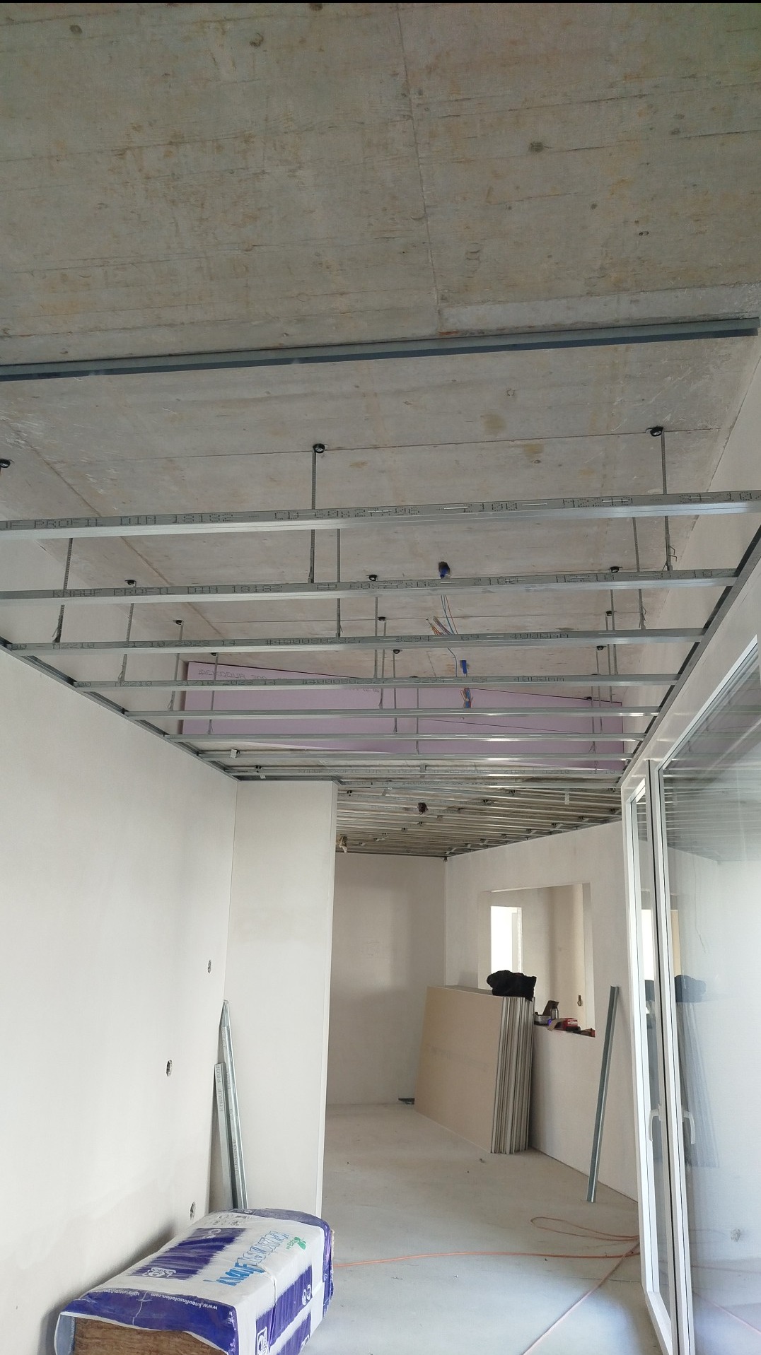 Montaż stelaża sufitu podwieszanego z płyt karton gips, widoczne profile, zawiesia i częściowo ułożona wełna mineralna, w tle ściany w stanie surowym i płyty gipsowe.