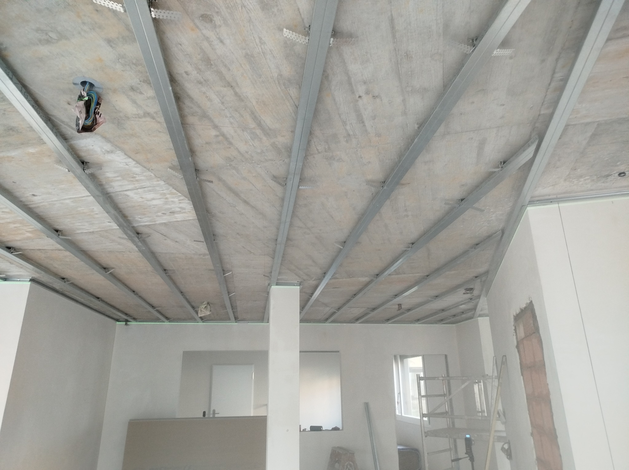 Montaż metalowej konstrukcji sufitu podwieszanego w surowym wnętrzu, widoczne kable elektryczne i laserowy poziom na ścianie.