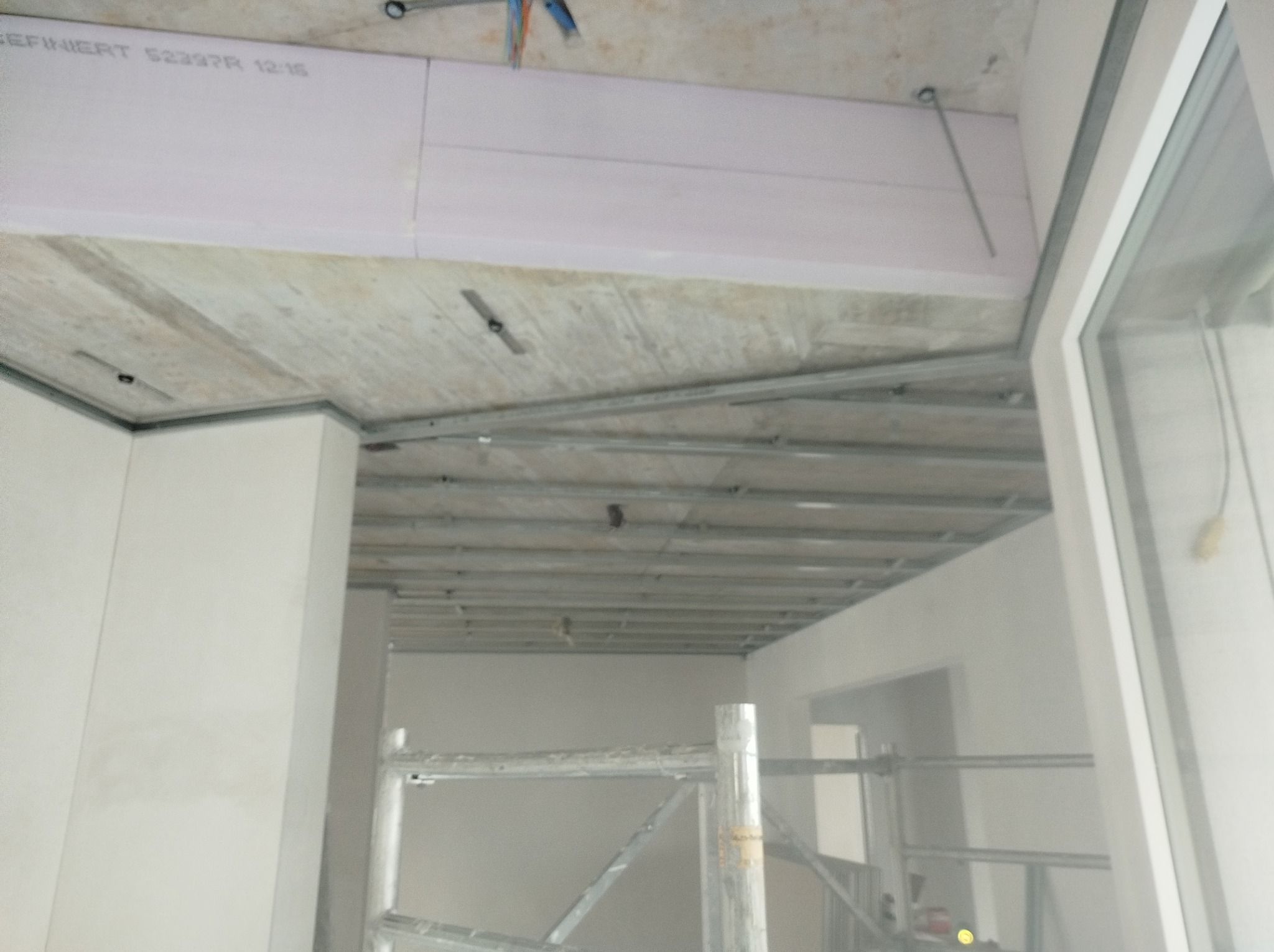 Montaż profili i płyt izolacyjnych na suficie podwieszanym, widoczne rusztowanie, surowy betonowy strop i instalacje elektryczne.