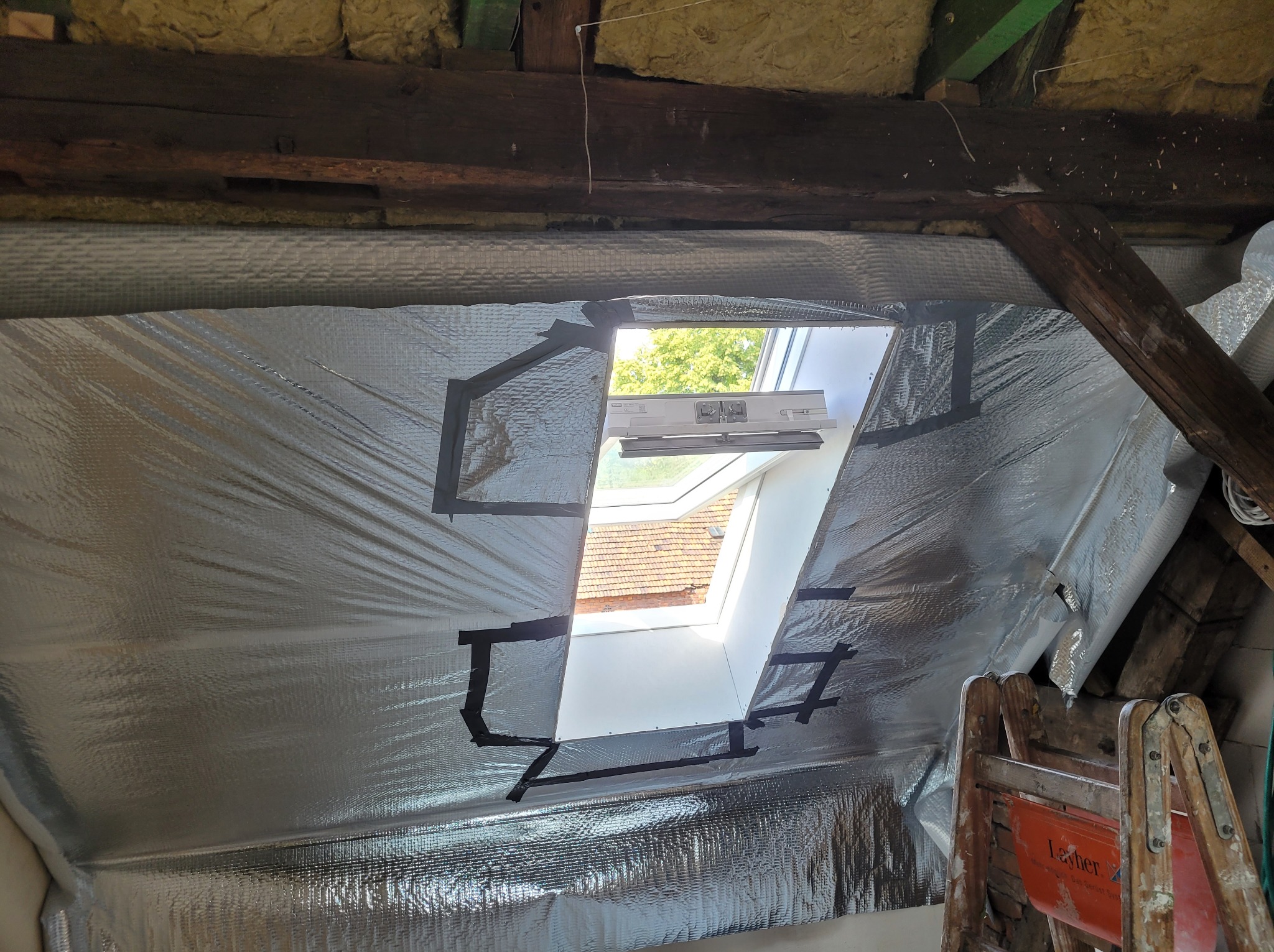 Folia termoizolacyjna zamocowana czarną taśmą wokół okna dachowego na poddaszu, widoczne drewniane belki konstrukcyjne i drabina.