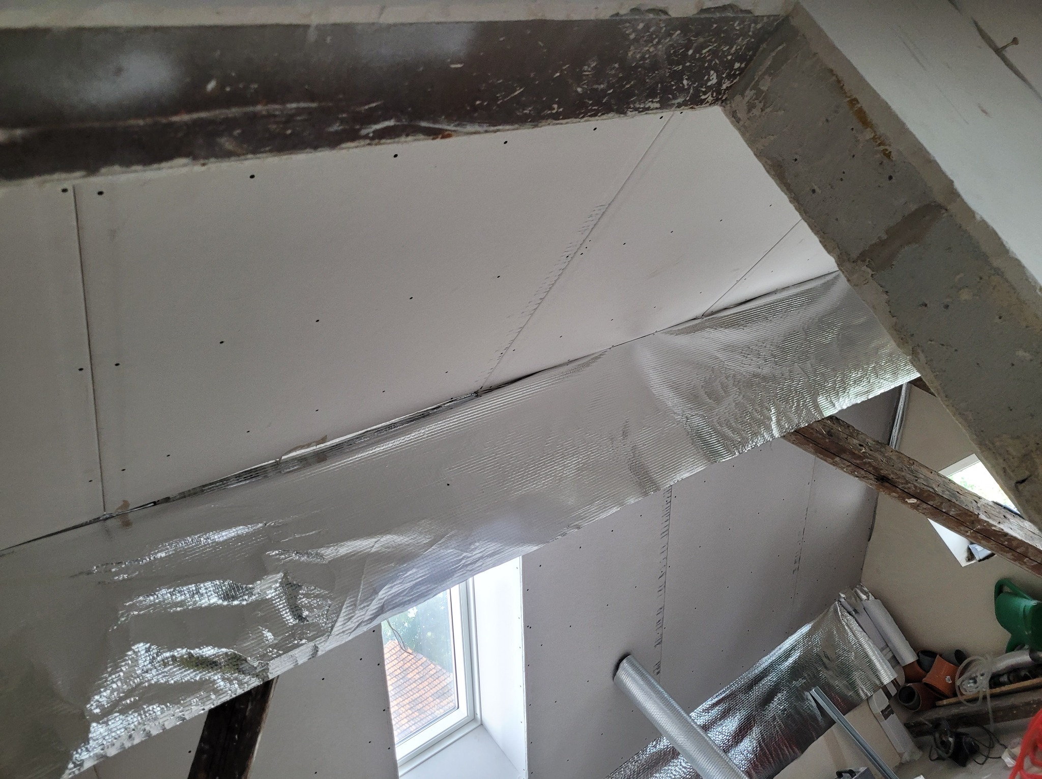 Skośny sufit na poddaszu w trakcie prac wykończeniowych, widoczne płyty kartonowo-gipsowe, izolacja folią aluminiową, drewniane elementy konstrukcyjne oraz okno dachowe.