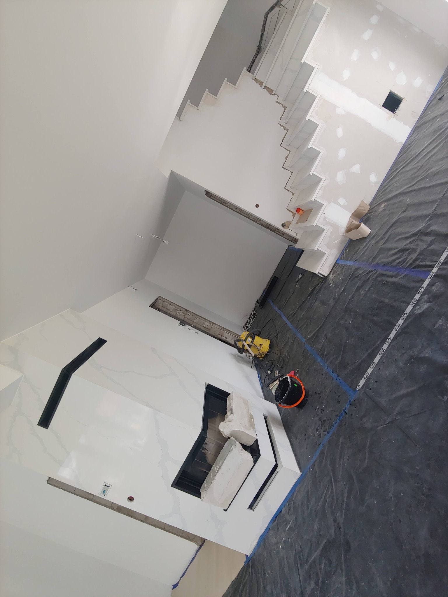 Wnętrze w trakcie prac wykończeniowych: białe ściany z marmurowym wzorem, czarny wkład kominkowy, surowe schody, podłoga zabezpieczona folią malarską, widoczny agregat i wiadro.