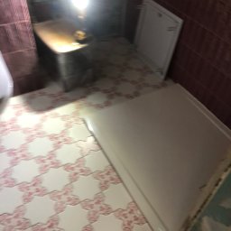 Renovation Hause Wojciech Tworkowski - Niedokończona łazienka z białą kabiną prysznicową, wzorzystymi płytkami podłogowymi w odcieniach bieli i różu, oraz fragmentem ściany w bordowych płytkach, oświetlona roboczą lampą.