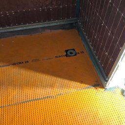 Renovation Hause Wojciech Tworkowski - Przygotowanie podłoża pod płytki w kabinie prysznicowej: pomarańczowa mata izolacyjna Schluter-DITRA 25 na podłodze, brązowe płytki ścienne w trakcie układania z krzyżykami dystansowymi.